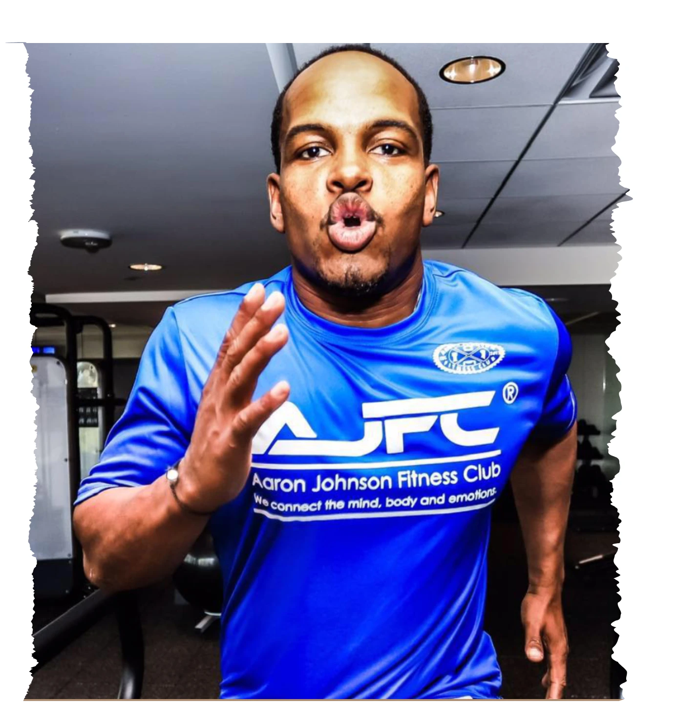 Online strength coach demonstrating a high-intensity running drill in a gym, wearing Aaron Johnson Fitness Club apparel