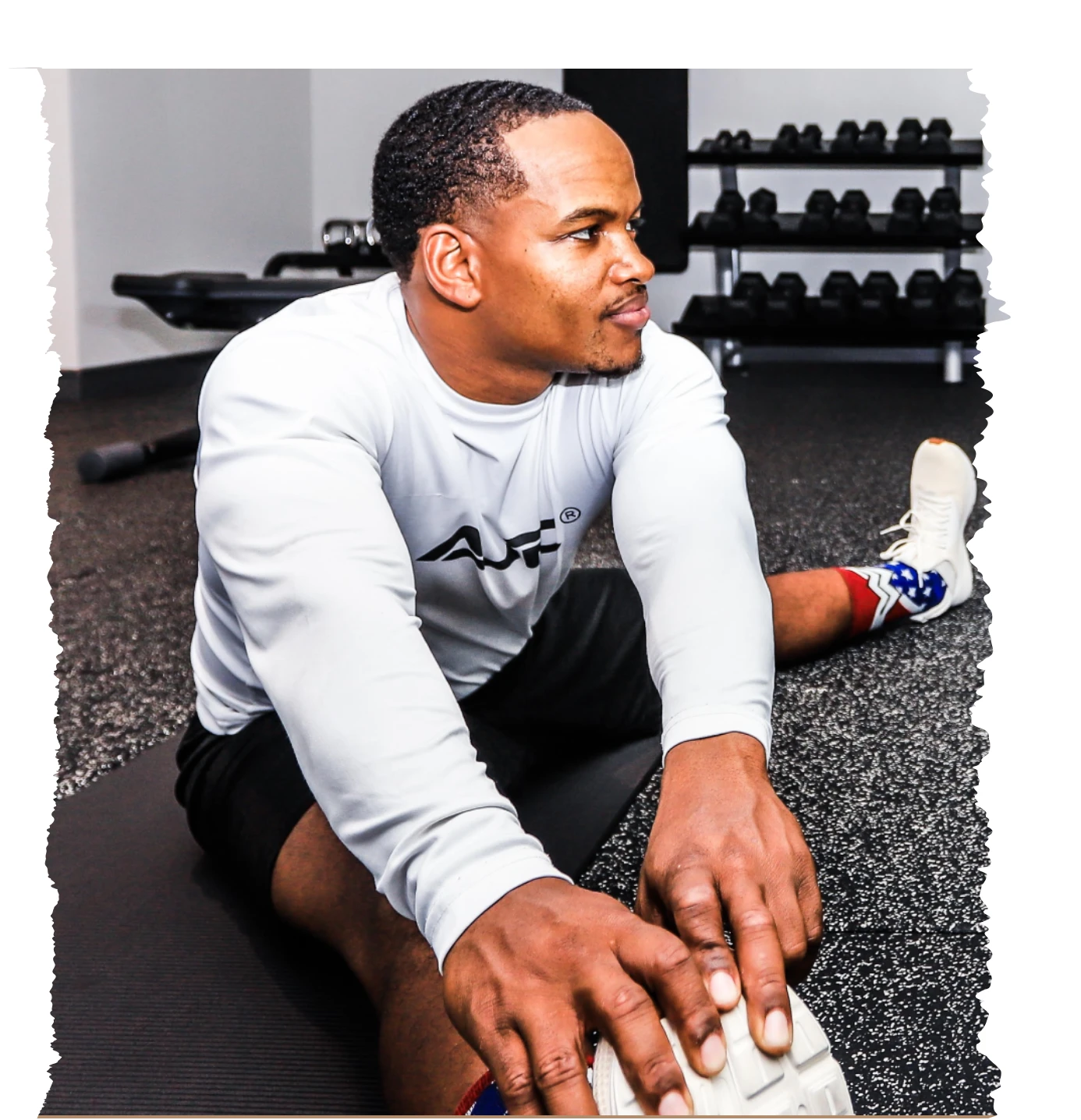 Online strength coach demonstrating a ham string stretch in a gym, wearing Aaron Johnson Fitness Club apparel.